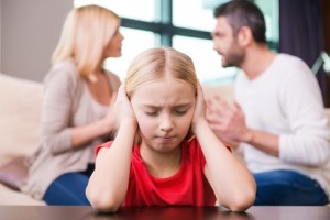 Please stop fighting! Depressed little girl leaning at the table and covering ear with hands while her parents shouting at each other in the background